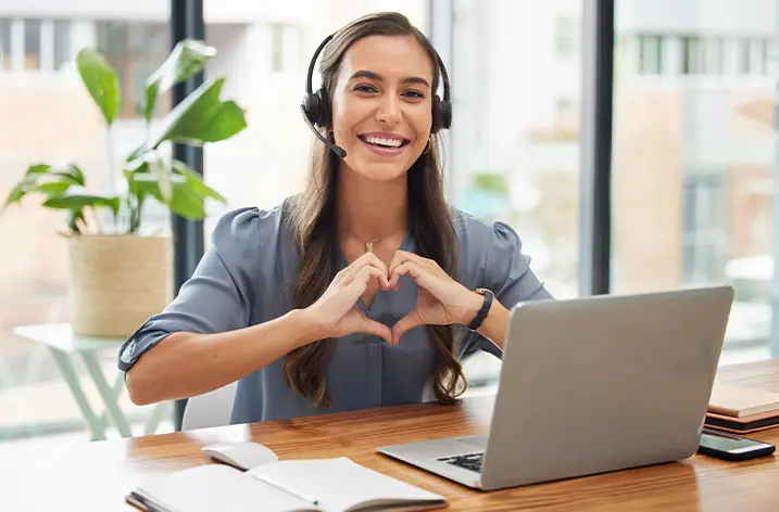 Mitarbeiterin mit Headset formt mit den Händen ein Herz vor dem Notebook