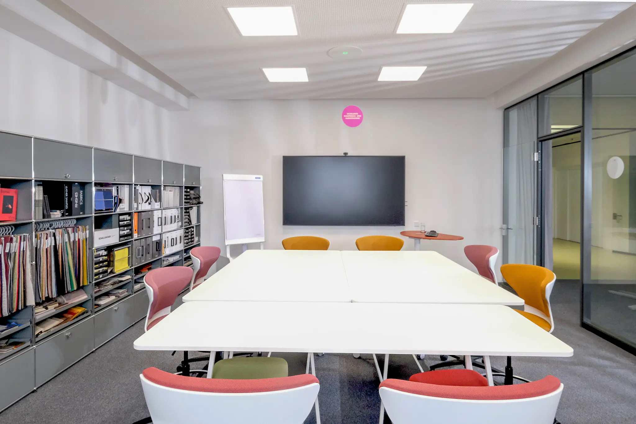 Besprechungsraum mit weißer Tafel, bunten Stühlen, Bildschirm und Musterregal