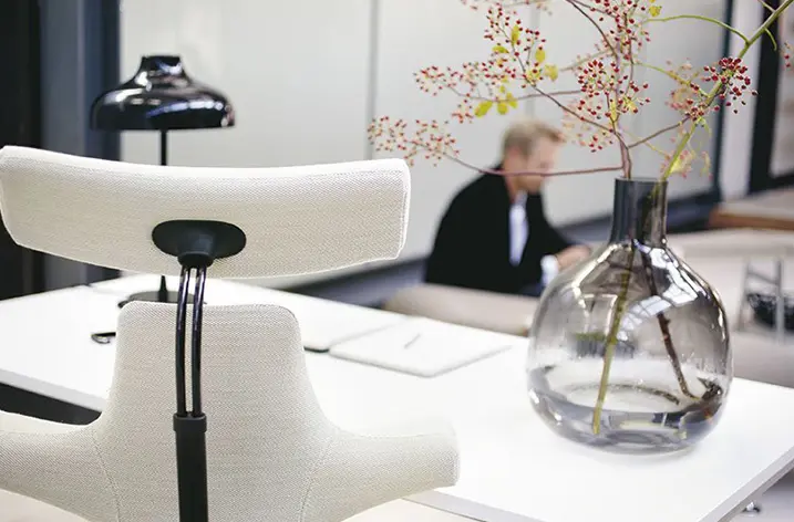 Weißer Sattelsitz-Drehstuhl am Schreibtisch, dahinter eine Vase mit Zweigen