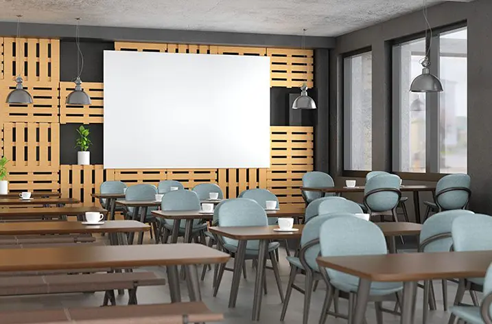 Kantine mit Holztischen, blaugrauen Stühlen und Leinwand vor einer Palettenwand