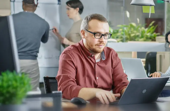 Mann arbeitet am Notebook im Großraumbüro, dahinter Kollegen am Whiteboard