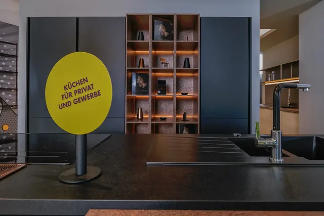 Küchenzeile im Showroom mit dunkler Arbeitsplatte und gelbem Schild „Küchen für privat und gewerbe“