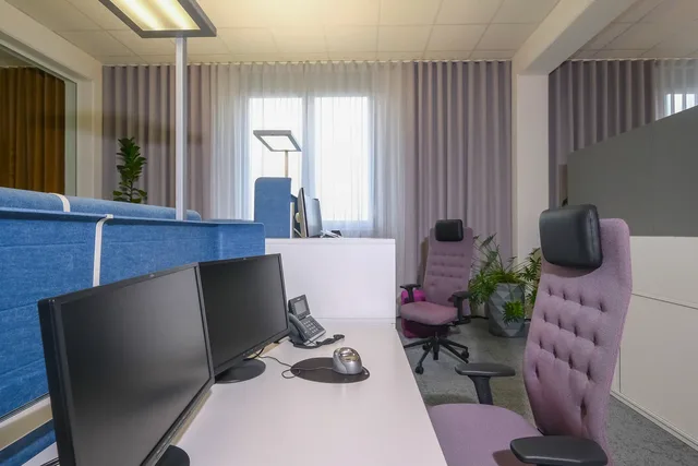 Arbeitsplätze mit blauen Trennwänden, dahinter altrosa Drehstühle am Fenster