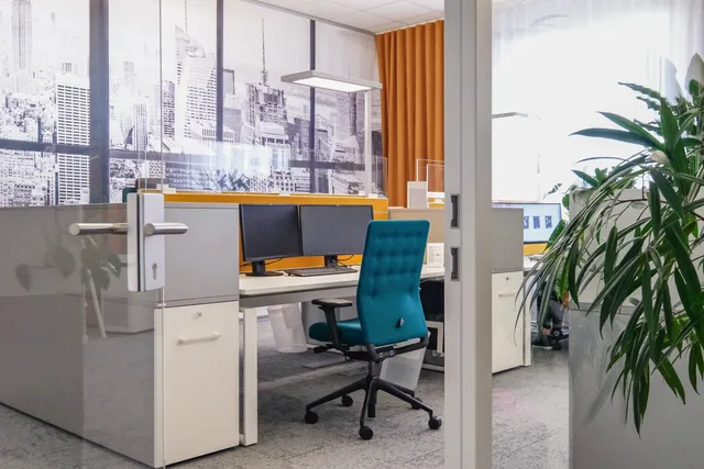 Blick durch die Glastür auf einen Arbeitsplatz mit türkisfarbenem Drehstuhl