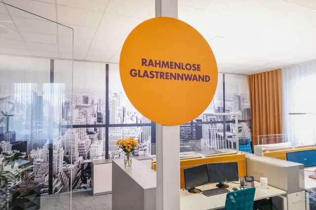 Orangefarbenes Schild „Rahmenlose Glastrennwand“ vor dem Großraumbüro mit Skyline-Fototapete