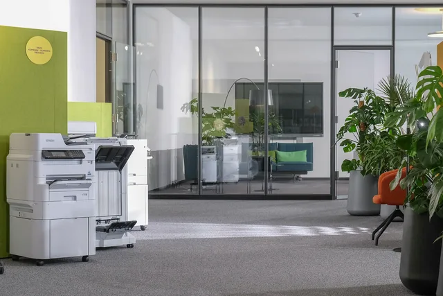 Multifunktionsdrucker vor grüner Akustikwand, dahinter verglaste Büroräume